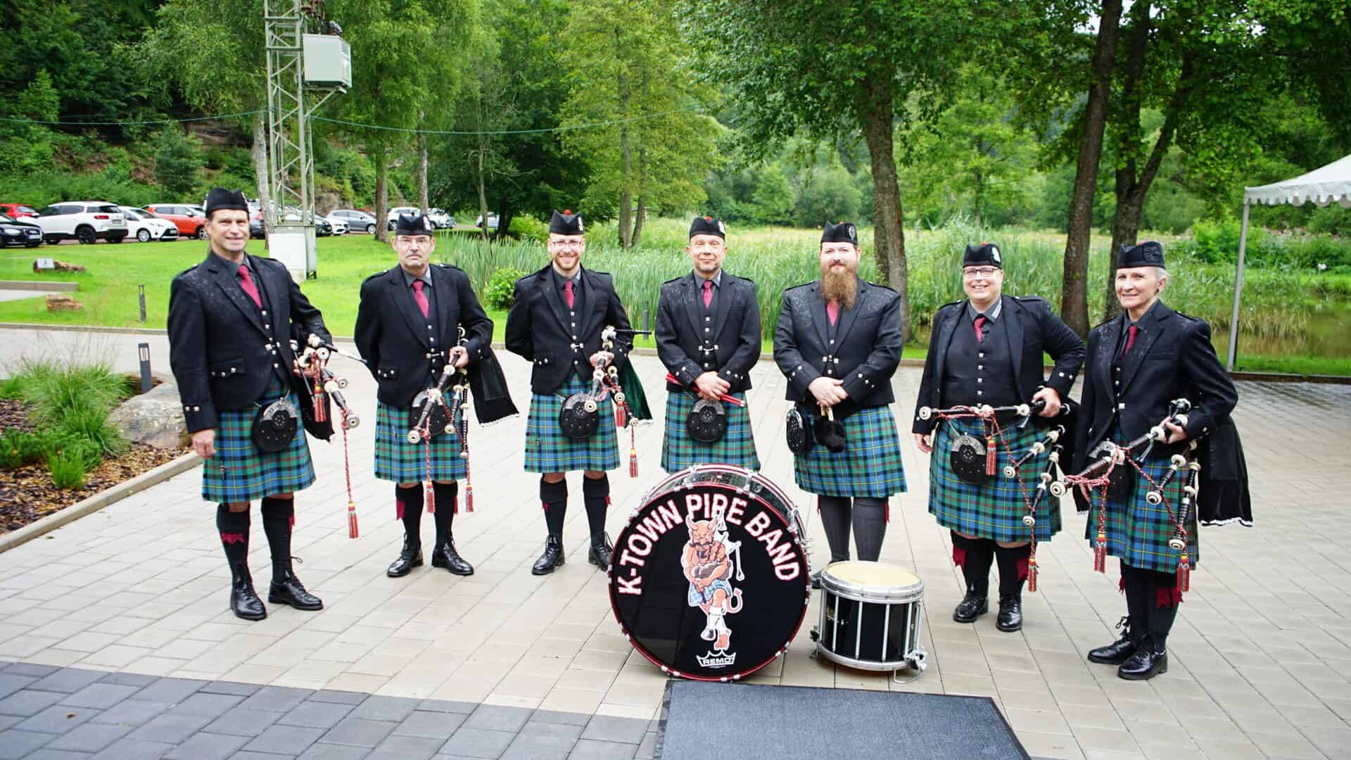 K-Town Pipe Band in traditioneller Kleidung bei einem Auftritt im Freien, mit Dudelsäcken und Trommeln, umgeben von Natur und Bäumen.